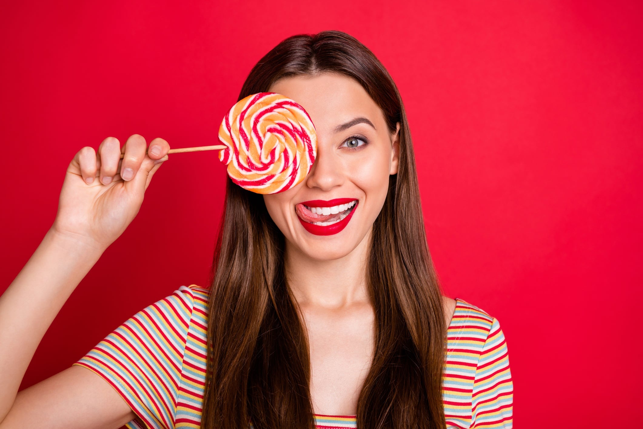 Image of woman holding, large lollipop over one eye and sticking tongue out. Red background.