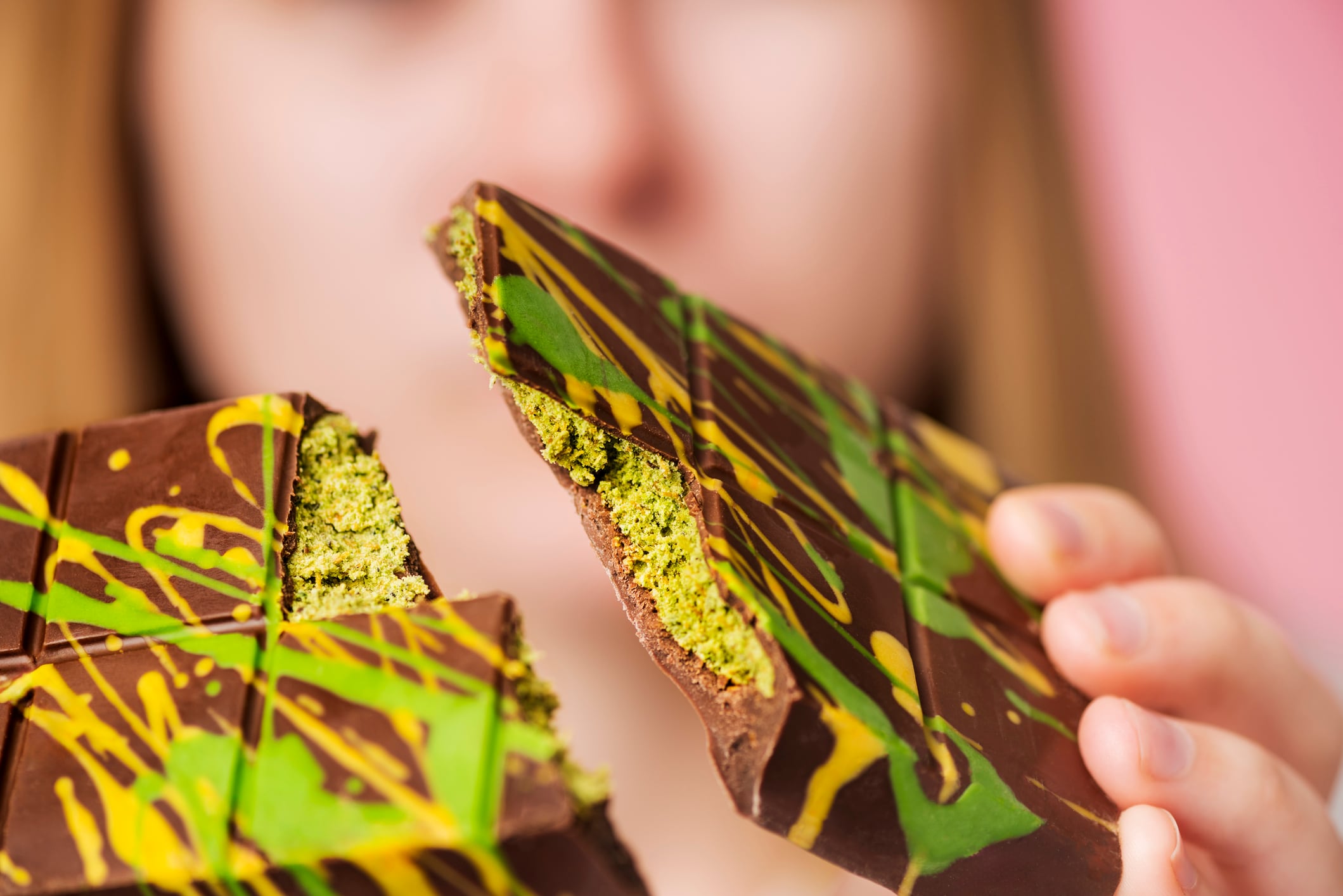 A young woman holds a bar of Dubai Chocolate that has been snapped in half towards the camera. The chocolate bar features random splashes of green and yellow, representing the key ingredients of pistachio cream and kataifi pastry. A shallow focus technique has been used to focus on the chocolate bar in the foreground.