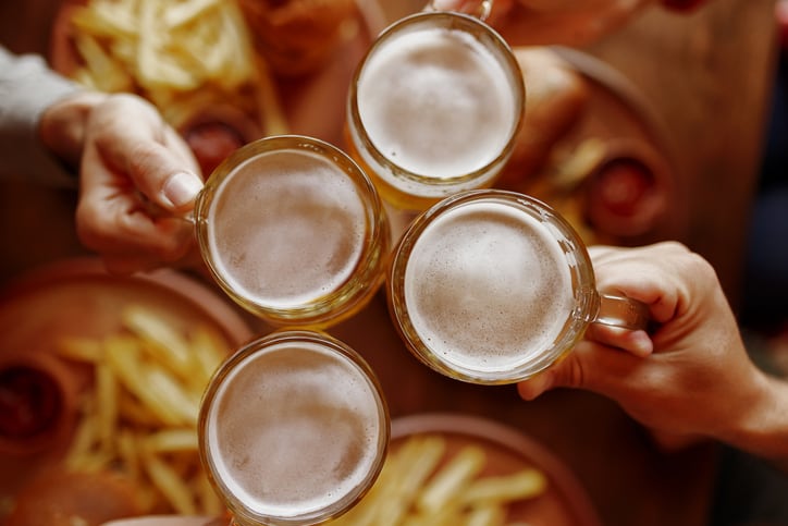 High-angle photo of beer glasses clinking with visible pub snacks. Leisure, celebration, food and beverage, friendly gathering.