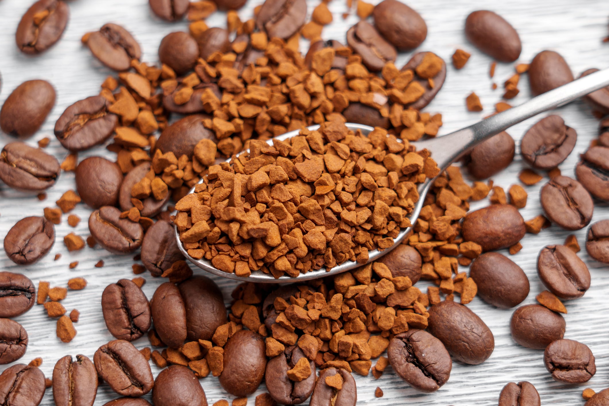 Soluble granules in metal spoon with scattered roasted Arabica coffee beans on aged white wooden surface. Mixed ingredients of instant and brewed beverage. Top View