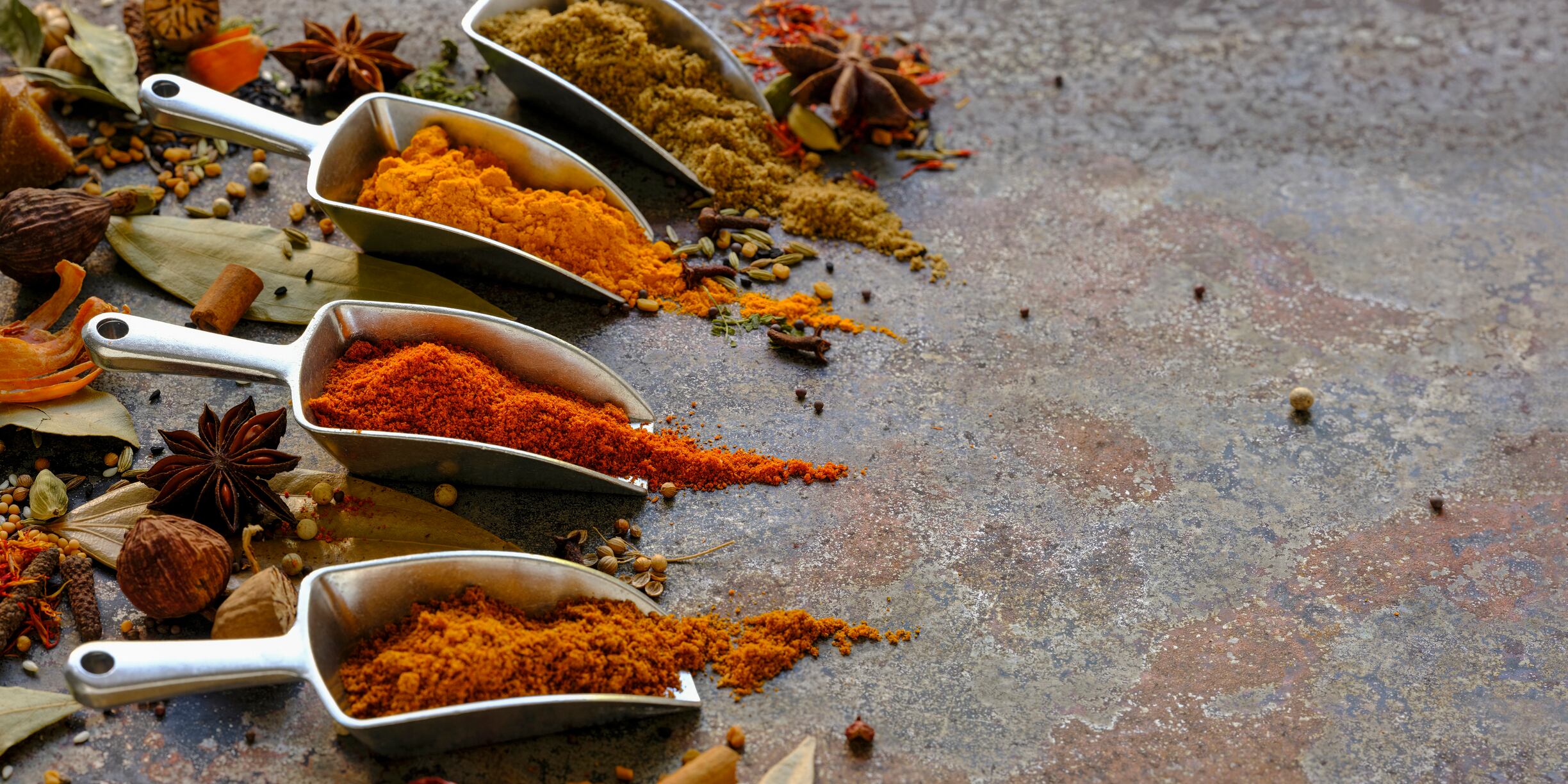 Many colorful, organic, dried, vibrant Indian food, ingredient spices in small aluminum metal spice scoops are arranged in a line on an old, weathered, rusted metal plate background. Taken from a high angle.