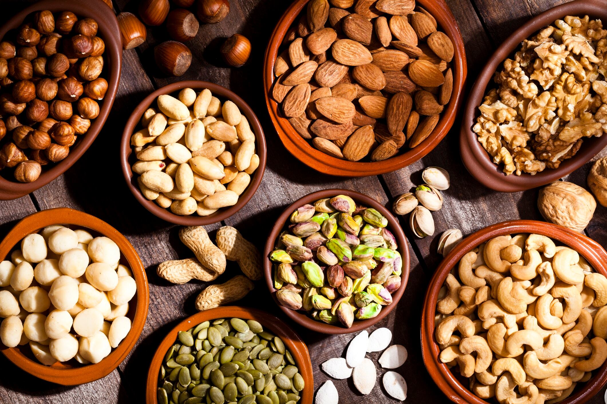 Top view of a rustic wood table filled with a large assortment of nuts like pistachios, hazelnut, pine nut, almonds, pumpkin seeds, peanuts, cashew and walnuts. Some nuts are in brown bowls and others are placed directly on the table.