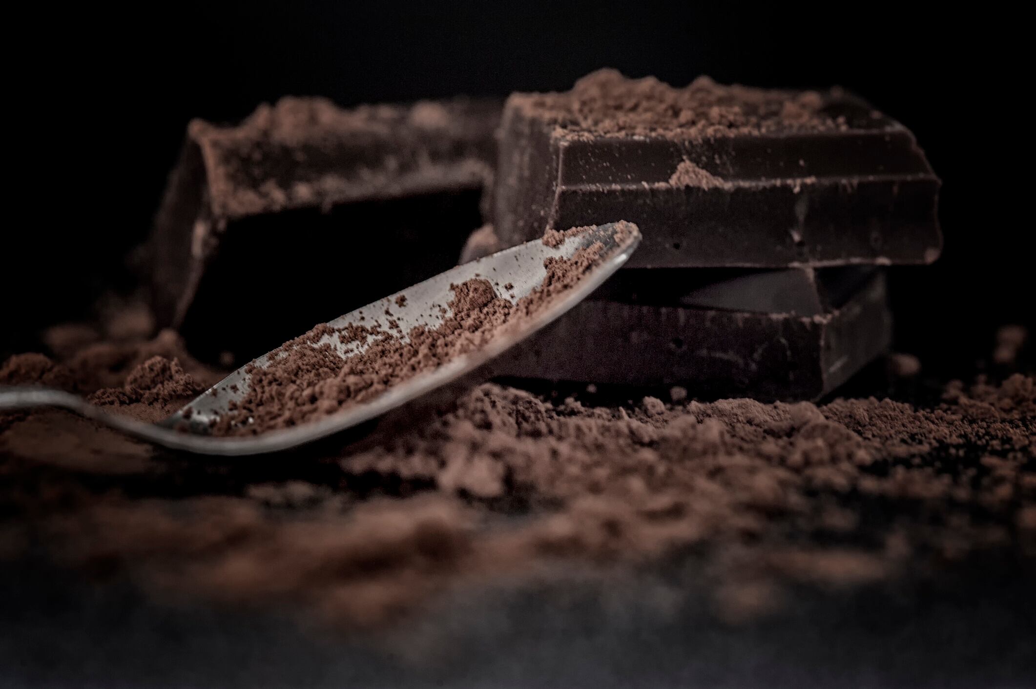 Close-up macro shot of chocolate pieces stacked on top of each other with sparse cocoa and spoon on black background.